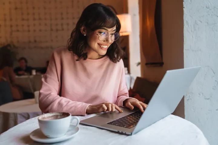 Mulher jovem em uma cafeteria, com notebook aberto e uma xícara de café ao lado, sorrindo para a tela.